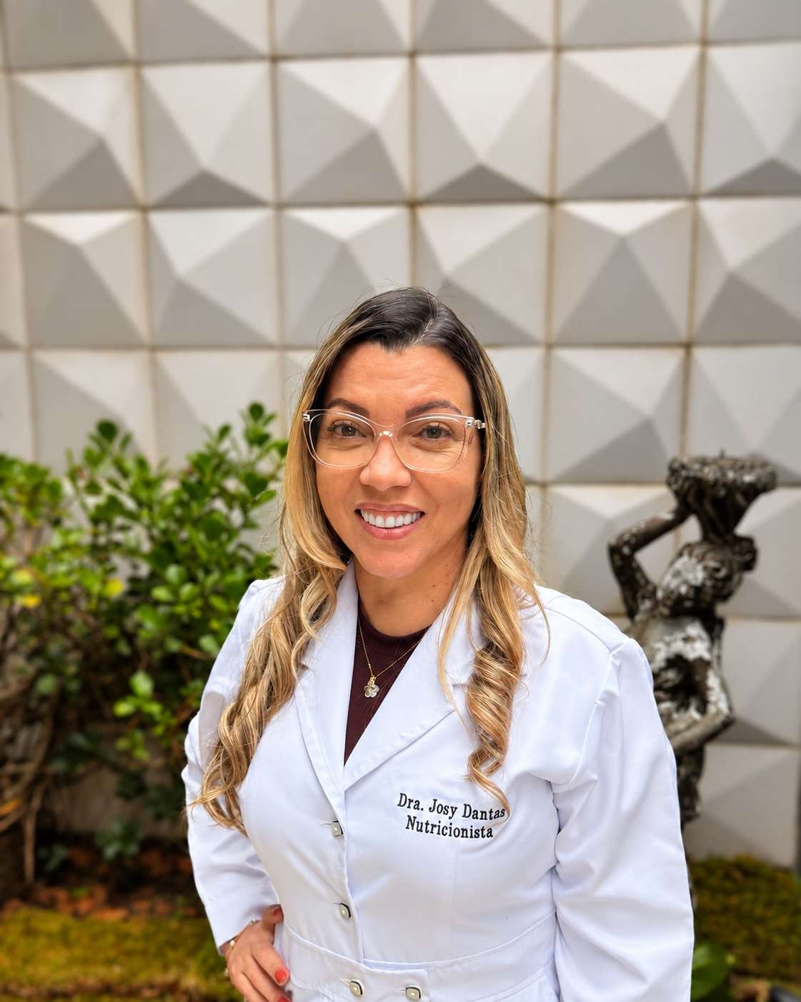 Dra. Josy Dantas, nutricionista, de jaleco branco, sorrindo em frente a um jardim